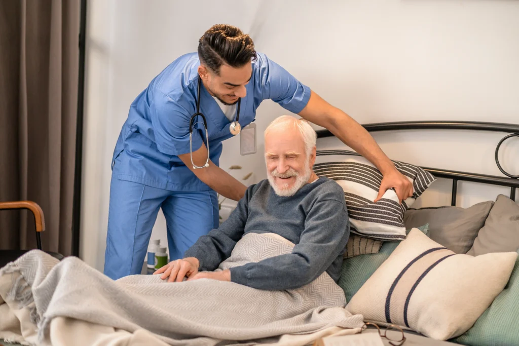 Un asistent medical în uniformă albastră și cu stetoscop ajută un bărbat în vârstă cu păr și barbă albă care stă în pat. Pacientul zâmbește în timp ce îngrijitorul îl susține, aranjând pernele.